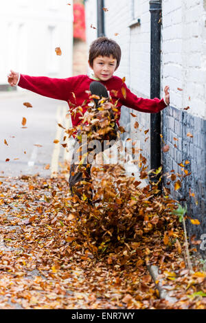 Bambino caucasico, ragazzo, 8-9 anni, camminando lungo una strada coperta di uscita verso lo spettatore, calciando le foglie in alto in aria. Autunno. Foto Stock