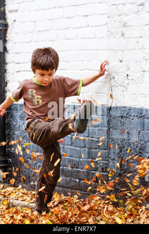 Bambino caucasico, ragazzo, 8-9 anni, camminando lungo una strada coperta di uscita verso lo spettatore, calciando le foglie in alto in aria. Autunno. Foto Stock