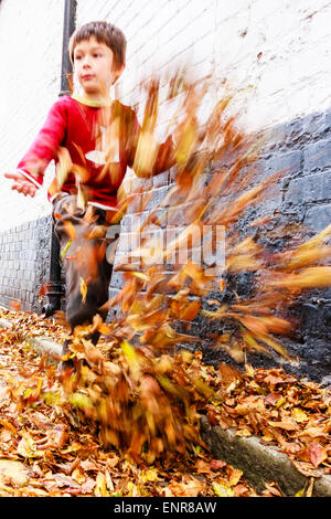 Bambino caucasico, ragazzo, 8-9 anni, camminando lungo una strada coperta di uscita verso lo spettatore, calciando le foglie in alto in aria. Autunno. Foto Stock