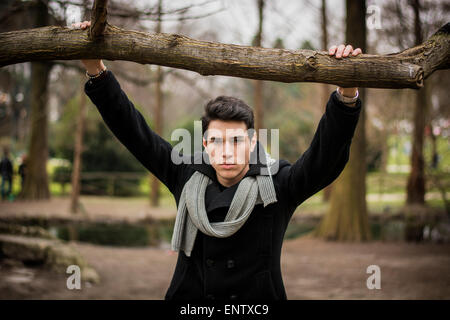 Bello elegante giovane uomo in nero elegante nero Cappotto invernale con sciarpa appoggiata sul ramo di albero mentre cercano per la fotocamera Foto Stock