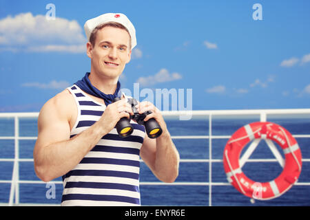 Giovane marinaio tenendo il binocolo e in piedi su un ponte di una barca con il blu del cielo e del mare in background Foto Stock