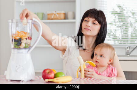 Carino brunette donna stacca una banana mentre si tiene il suo bambino sulle ginocchia in cucina Foto Stock