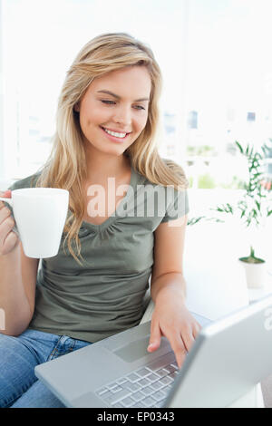 Donna sorridente come usa il suo computer portatile e contiene una tazza nella sua mano libera Foto Stock