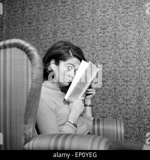 Norwegische Schlagersängerin Wencke Myhre versteckt sich im Wohnzimmersessel hinter einem Buch, 1960er Jahre. Schl norvegese Foto Stock