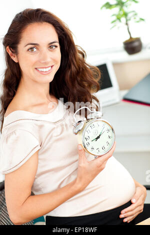 Gravidanza radiante imprenditrice tenendo una sveglia e seduto alla sua scrivania nel suo ufficio Foto Stock