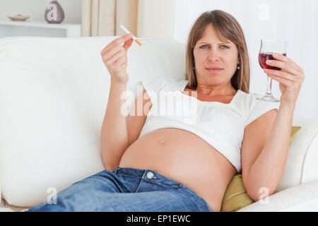 Incantevole donna incinta tenendo una sigaretta e un bicchiere di vino rosso mentre giaceva su un divano Foto Stock