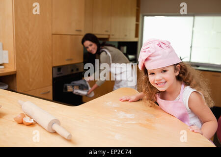 Ragazza seduta in cucina mentre sua madre mette i cookie nel forno Foto Stock