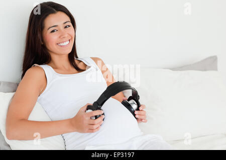 Vista frontale di un attraente donna incinta mettendo le cuffie sul suo ventre mentre giaceva su un letto Foto Stock