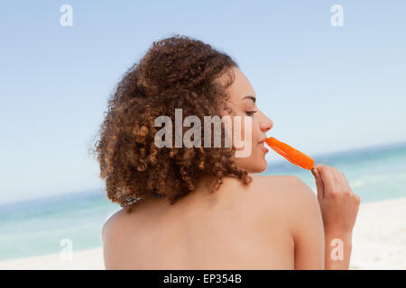 Attraente giovane brunette mangiare un arancio per lecca-lecca di ghiaccio di fronte all'oceano Foto Stock