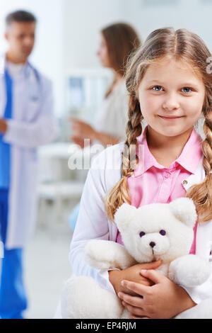 Carino il bambino con Orsacchiotto bianco guardando la fotocamera mentre il medico parla con sua madre sullo sfondo Foto Stock