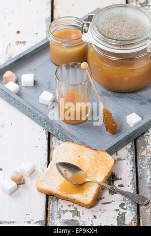 Toast con fatti in casa la salsa di caramello per la prima colazione, servita con il cucchiaio, vasetti e cubetti di zucchero sul vassoio grigio sopra il vecchio tavolo in legno Foto Stock