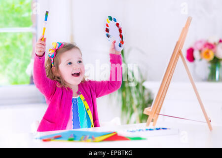 Carino felice bambina, adorabile preschooler, dipinto con colori ad acqua su tela in piedi su un cavalletto di legno in una stanza soleggiata Foto Stock