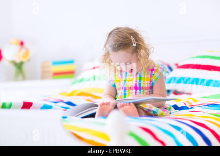 Ricci carino piccolo bimbo ragazza la lettura di un libro seduti in una luminosa camera da letto su un di legno letto bianco con colorate lenzuola arcobaleno Foto Stock