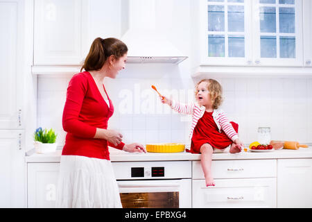 Giovane madre e la sua adorabile figlia, carino divertente toddler ragazza in un abito rosso, la cottura di una torta insieme a preparare il pranzo in cucina Foto Stock