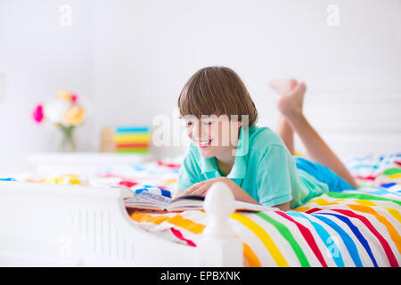 Sorridenti ragazzo scuola leggendo un libro rilassante su un bianco letto in legno con manto colorato in una stanza soleggiata Foto Stock