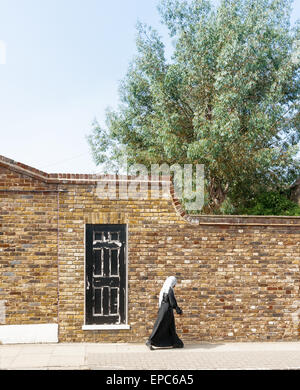 Donna musulmana in prua East London vestito di bianco e nero passa in bianco e nero porta nella parete di mattoni Foto Stock