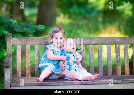 Adorabili bambini, poco ragazza di ricci e un simpatico baby boy, fratello e sorella, seduti insieme su una panca in legno in un giardino Foto Stock