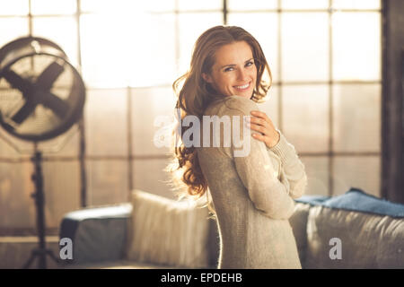 Un sorridente brunette donna in abbigliamento comodo è in piedi in un loft soggiorno, avvolgente se stessa mentre guardando sopra la sua spalla. Urban Chic loft dettagli decorativi. Foto Stock