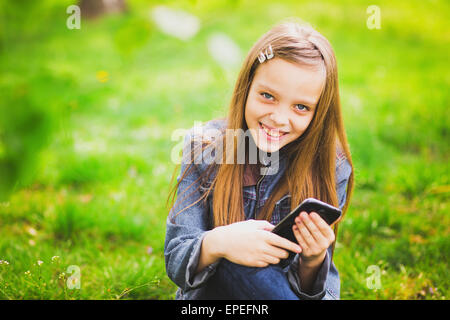 Ritratto di sorridente ragazza seduta nel parco e scrivere messaggi sms utilizzando il telefono cellulare Foto Stock