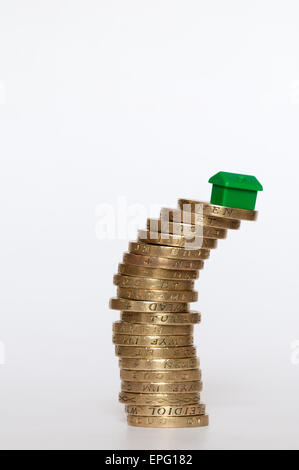 Un piccolo modello di plastica green house bilanciato sulla sommità di una pila di inclinazione di libbra di monete. Finanziamento di alloggi concetto. Foto Stock