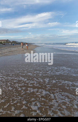 La marea schiumosa entra nella copertura della spiaggia di fronte la gente camminare lungo l'oceano sotto il cielo azzurro e luminoso puffy nuvole Foto Stock