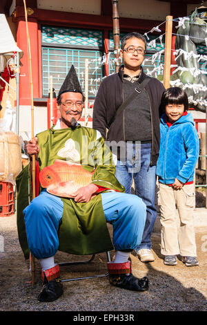 Un padre e un figlio sorridenti che si posano accanto ad un uomo seduto vestito da Dio del pescatore, Ebisu, al festival di nuovo anno al santuario di Shinto Nishinomiya. Foto Stock