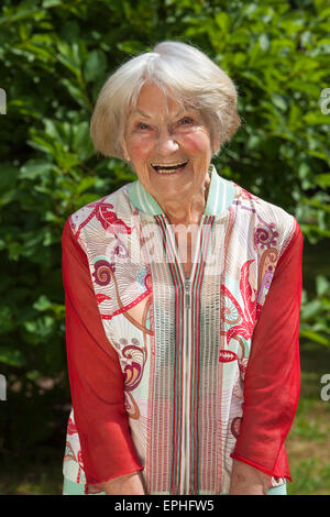 Ridendo attraente pensionato femminile in un elegante vestito rosso sorridente gioiosamente alla telecamera come essa si erge all'aperto in giardino Foto Stock