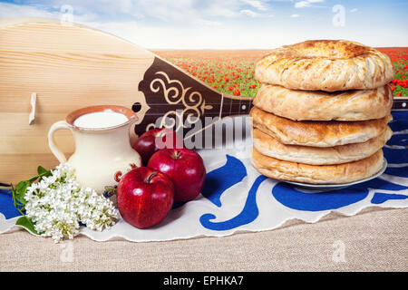 Dombra strumento kazako, mele, latte e pane lepeshka sul tavolo al fiore di papavero dello sfondo dei campi Foto Stock