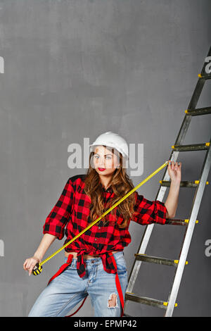 Affascinante ragazza builder tenendo un nastro di misurazione. Ragazza vestita in jeans, un plaid camicia e un casco. Spazio per il testo. Foto Stock