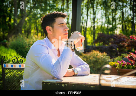 Bel giovane uomo seduto da solo al tavolo esterno in posizione di parcheggio o di natura, con bicchiere di bevanda di soda Foto Stock