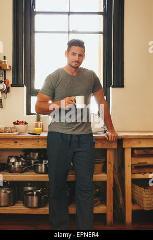 Ritratto di giovane uomo in piedi in cucina tenendo una tazza di caffè. Bel maschio modello avente fresca del mattino il caffè a casa in k Foto Stock