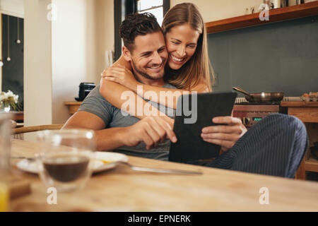 Coppia sorridente come leggere un computer tablet insieme nella mattina in cucina. Giovane uomo e donna recuperando sui social medi Foto Stock
