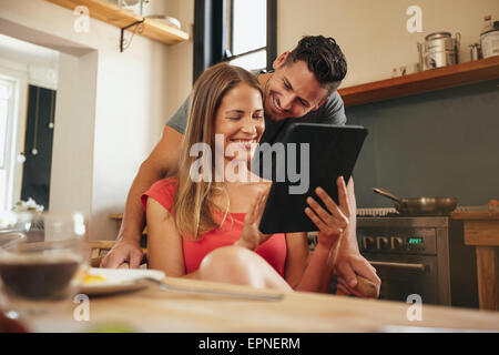 Coppia sorridente utilizzando un computer tablet insieme nella mattina in cucina. Giovane uomo e donna al tavolo per la colazione con digital tabl Foto Stock