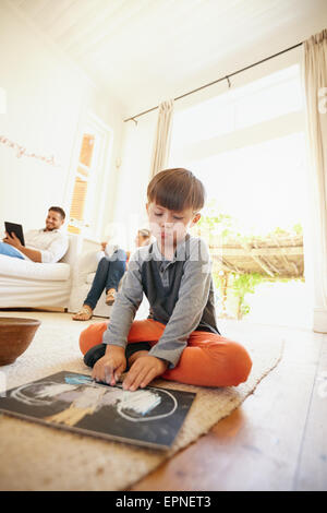 Piccolo scolaro seduto sul piano di disegno con i suoi genitori seduti in background sul lettino. Famiglia in soggiorno. Foto Stock