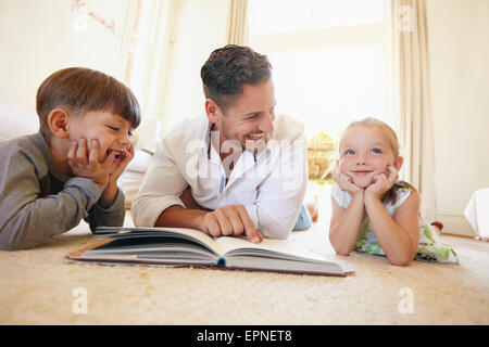 Ritratto di felice giovane famiglia di tre sdraiato sul pavimento con un libro. Padre di due bambini la lettura di un libro di storia in soggiorno Foto Stock