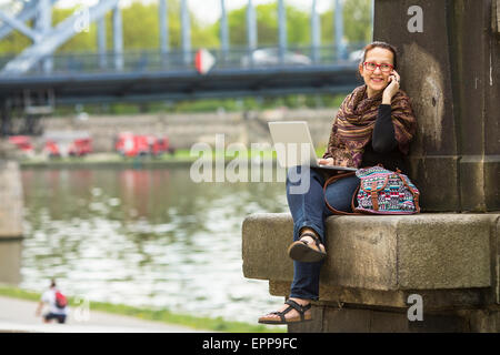 Giovane donna con il computer portatile seduti lungo gli argini del fiume nella città vecchia a parlare su un telefono cellulare. Foto Stock