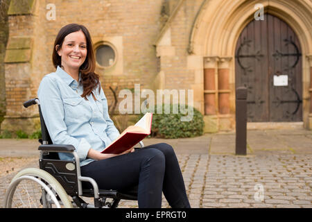 Giovane donna in una sedia a rotelle la lettura di una bibbia al di fuori di una chiesa Foto Stock