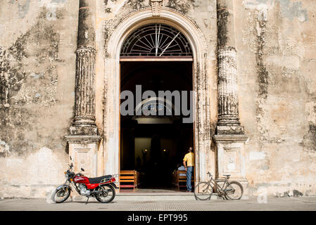Iglesia De La Merced Facciata Barocca Ingresso Granada Nicaragua | GRANADA, Nicaragua — Un parcheggio per motociclette e biciclette parcheggiato davanti all'ingresso barocco aperto dell'Iglesia de la Merced, colonne scanalate che incorniciano un'anteprima della navata e della sua vetrata oltre. L'ingresso principale sopravvive in gran parte da una ricostruzione del XVIII secolo, mentre l'interno visibile riflette la ricostruzione dopo che le forze di William Walker hanno bruciato la chiesa nel 1856. Foto Stock
