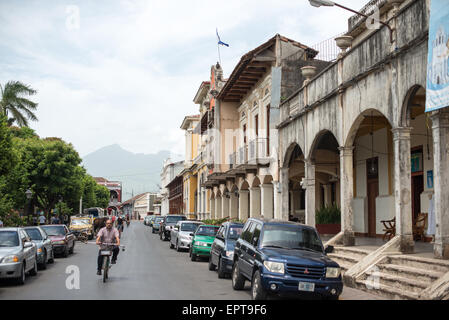 Architettura coloniale spagnola Parque Central Granada Nicaragua // GRANADA, Nicaragua - Una strada che confina con Parque Central mostra l'architettura coloniale spagnola conservata che definisce il quartiere storico di Granada. Parque Central è la piazza principale e il cuore culturale di Granada, una delle più antiche città coloniali del Nicaragua, fondata nel 1524. I colorati edifici coloniali che fiancheggiano la strada presentano elementi caratteristici tra cui facciate ornate, balconi in legno e cortili interni tipici del design coloniale spagnolo. Le montagne sorgono in lontananza oltre la città, parte del Foto Stock