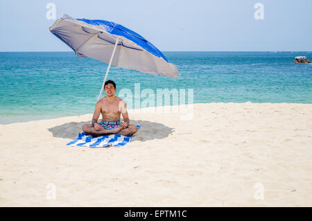 Maschio atletico yoga in posizione del loto di sedersi su un asciugamano sotto blu e grigio ombrellone sulla spiaggia con l'oceano sullo sfondo Foto Stock