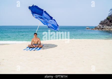 Maschio atletico yoga in posizione del loto di sedersi su un asciugamano sotto blu e grigio ombrellone sulla spiaggia con l'oceano sullo sfondo Foto Stock