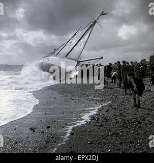 Degli anni Cinquanta, storico, una barca a vela capovolta o inginocchiato sul suo lato su una spiaggia sassosa, con grande folla che guarda. Foto Stock