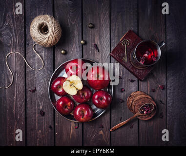 Le mele rosse sulla piastra e la tazza con il tè di ibisco sul vecchio libro al buio sullo sfondo di legno Foto Stock