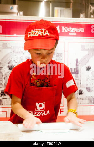 Giappone, Nishinomiya, Kidzania. Western boy in rosso grembiule, pasta di rotolamento con una mano rendendo la pizza. Vista frontale senza contatto visivo. Foto Stock