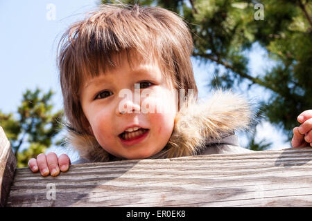 Colpo di testa e di spalla di bambino, ragazzo, 3-4 anni, con guance rosse, con cappotto invernale con colletto di furry, guarda su una recinzione di legno presso lo spettatore. Foto Stock