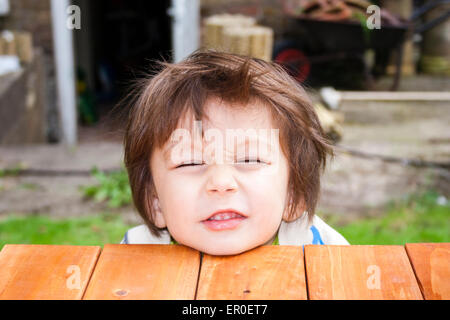 Primo piano del viso del bambino, ragazzo, 3-4 anni, mentre si appoggia il mento su una piattaforma di legno, di fronte allo spettatore, e svitando il volto verso l'alto. All'aperto Foto Stock