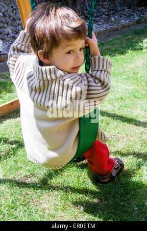 Bambino piccolo, ragazzo, 3-4 anni, seduto su un'altalena da giardino. Visto da dietro mentre guarda indietro sulla spalla verso lo spettatore. Indossa un cardigan spesso. Foto Stock