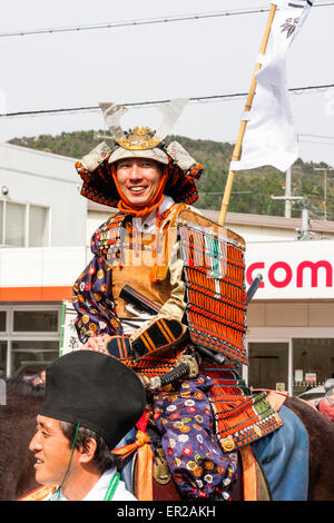 Uomo giapponese vestito con armatura samurai completa a cavallo sorridendo allo spettatore mentre si attraversa la strada della città nella sfilata Genji a Tada in Giappone. Foto Stock