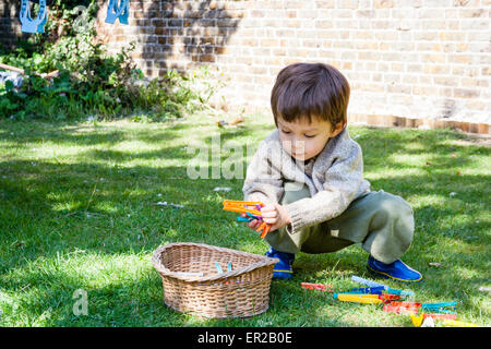 Bambino, ragazzo, 4-5 anni, alla luce del sole, accovacciato sul prato in giardino mentre raccoglie i vestiti pioli e li mette in un cesto di vimini. Foto Stock