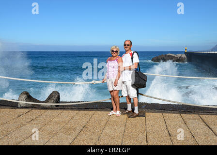 Coppia conciate giovane in abiti leggeri è in piedi sul terrapieno in luce del sole su un blu brillante acqua e cielo e spruzzi wave indietro Foto Stock